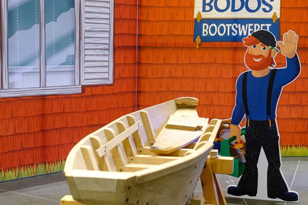 Ein Ausstellungsstück einer Bootswerft mit einem hölzernen Boot im Bau und einer Cartoon-Figur eines Bootsbauers, um eine interaktive und lernorientierte Umgebung für Besucher darzustellen.