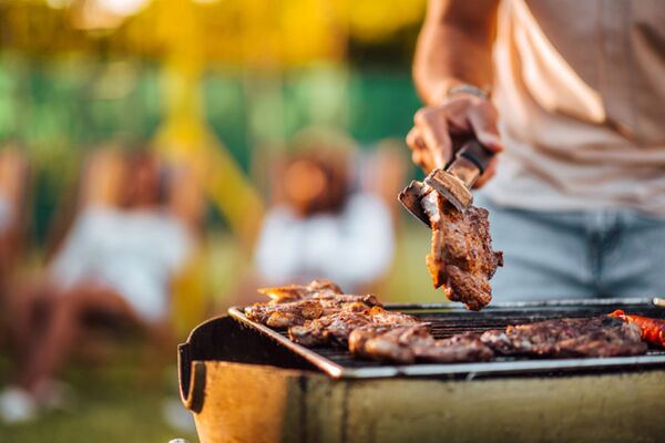 Ein Barbecue mit einer Person, die saftige Fleischstücke auf einem Grill wendet, während Menschen im Hintergrund entspannen.
