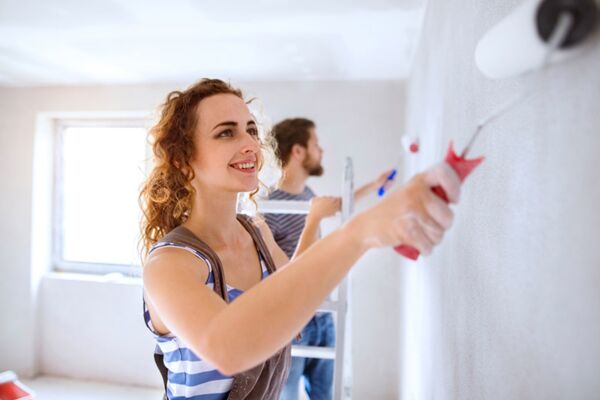 Eine junge Frau lacht, während sie fröhlich die Wände ihrer neuen Wohnung streicht, umgeben von Freunden oder Familie.