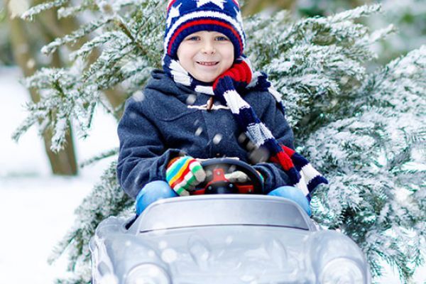Ein glückliches Kind fährt in einem kleinen Auto durch eine verschneite Landschaft, mit einem Weihnachtsbaum.