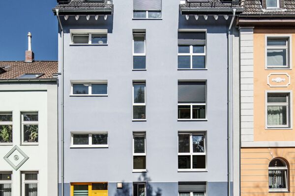 Das Bild zeigt die Fassade eines frisch renovierten Mehrfamilienhauses, eingebettet zwischen traditioneller Architektur. Die klaren, modernen Linien und die neutralen Farbtöne der Fassade heben das Gebäude von seinen farbenfroheren Nachbarn ab.