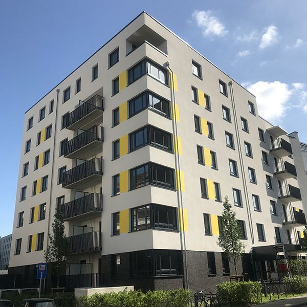 Ein neues Apartmentgebäude mit einem auffälligen gelben und grauen Farbschema. Das Gebäude bietet Balkone und eine klare, blockhafte Struktur, die urbanes Wohnen in einer modernen Umgebung widerspiegelt.