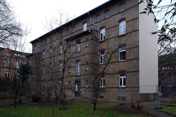 Historisches Mehrfamilienhaus aus Ziegelsteinen mit großen Fenstern, umgeben von Bäumen in einer städtischen Umgebung.