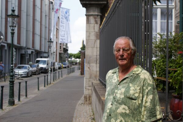 Ein älterer Mann in einem lockeren Hemd steht in einer historischen Stadtstraße, flankiert von klassischen Laternen und moderner Architektur.