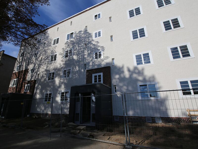 Fotografie eines frisch renovierten Wohngebäudes mit Schatten von Bäumen auf der weißen Fassade und Schutzzaun vor dem Eingang.