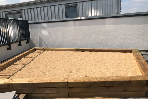 Auf einem Dach angelegter Sandspielbereich für Kinder, umgeben von einem niedrigen Holzzaun.