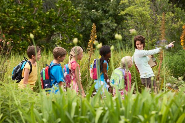 Eine Lehrerin oder Betreuerin führt eine Gruppe von Kindern mit Rucksäcken durch eine grüne, bewachsene Landschaft.