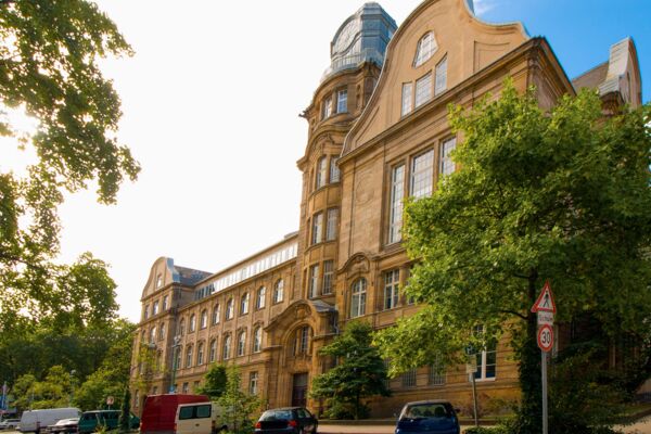 Ein beeindruckendes, altes Gebäude mit architektonischen Details, das bei Tageslicht vor einer grünen Kulisse glänzt.