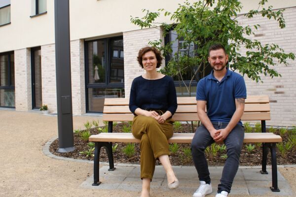 Zwei Personen sitzen auf einer Parkbank in einer urbanen, begrünten Umgebung vor einem modernen Gebäude und lächeln.