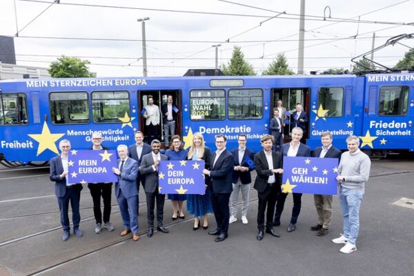 Politiker und Bürger posieren vor einer Straßenbahn, die für die Europawahl 2024 wirbt. Eine Aktion, die die Bedeutung der Wahl und europäischer Werte unterstreicht.