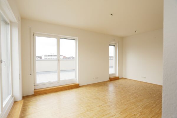 Ein gut beleuchtetes Zimmer mit polierten Holzböden und weitreichenden Fenstern, die auf einen privaten Balkon führen, bietet einen frischen, luftigen Wohnraum.