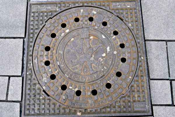 Nahaufnahme eines Schachtdeckels mit städtischem Wappen, was auf kommunale Dienstleistungen und Infrastruktur hinweist.