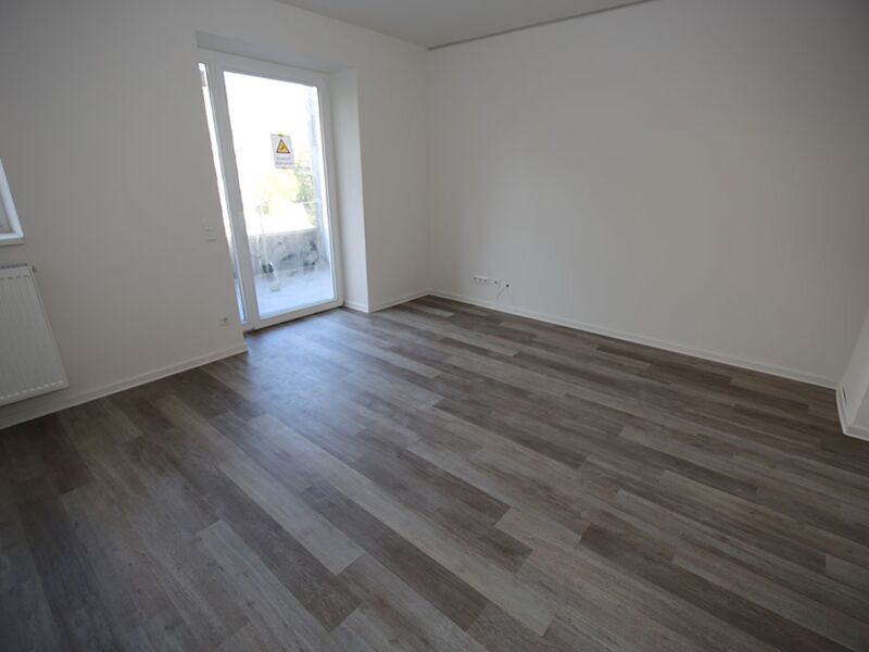 Innenraumansicht eines leeren Zimmers mit Holz-Laminatboden und Terrassentür, die zu einem kleinen Balkon führt.