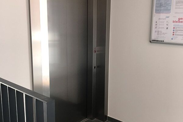 Moderner Aufzug in einem Wohngebäude, eingefasst in einer Metalltür mit Aushängen zur Sicherheit und Information.