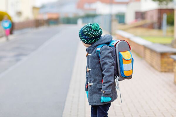 Bild eines kleinen Kindes von hinten betrachtet, das mit einer bunten Schultasche auf einer leeren Straße geht, symbolisch für den Beginn eines neuen Schuljahres oder Schultages.