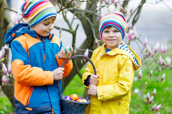 Ein fröhliches Bild von zwei Kindern mit bunten Ostereiern in einem Korb, die im Frühling unter blühenden Bäumen spielen, was die Freude der Feiertage hervorhebt.