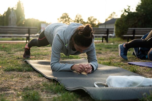 Eine junge Frau führt auf einer Matte im Park eine Plank-Übung durch, umgeben von anderen Teilnehmern in einem städtischen Fitnesskurs bei Sonnenuntergang.