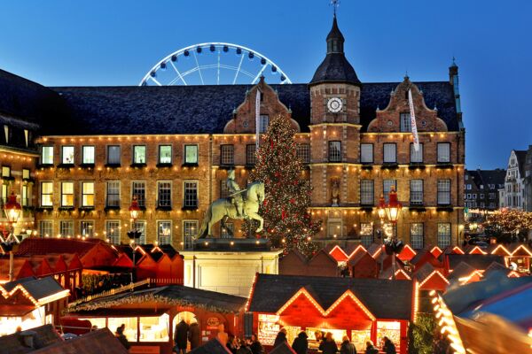 Ein lebendiges Bild eines Weihnachtsmarktes in der Dämmerung vor einem prächtig beleuchteten historischen Rathaus, mit einem großen Weihnachtsbaum und einem Riesenrad im Hintergrund, was eine festliche und fröhlic