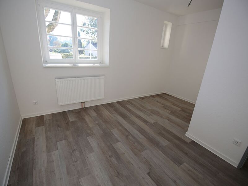 Bild eines leeren, gut beleuchteten Zimmers mit großem Fenster und Holzboden.