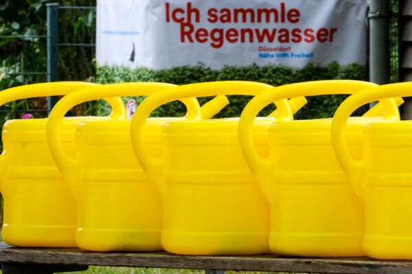 Eine Reihe leuchtend gelber Gießkannen, die die Bedeutung der Regenwassernutzung betonen, unterstützt durch eine lokale Umweltkampagne.