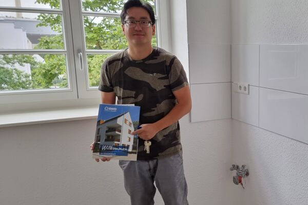 Ein Mann, der stolz den Schlüssel zu seiner neuen Wohnung hält, neben einer Broschüre mit der Aufschrift „Willkommen“.
