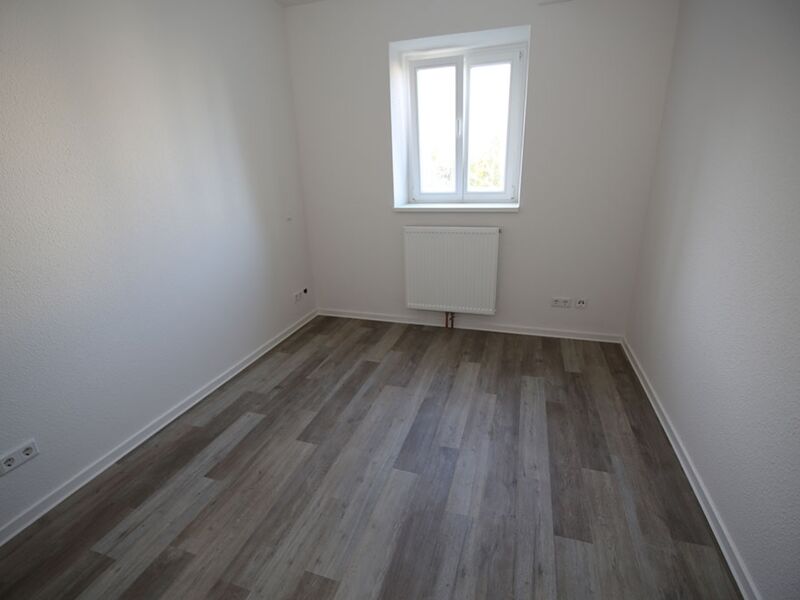 Foto eines weißen, leeren Raumes mit einem Holzboden und einem Fenster, das viel Tageslicht hereinlässt.