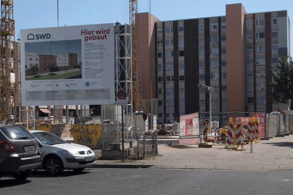 Ein Bauschild vor einer Baustelle mit Wohnblöcken im Hintergrund, das Informationen über das Bauvorhaben und den Bauträger bietet.