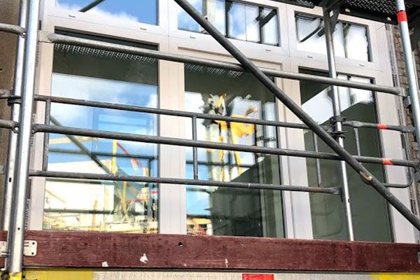Baustelle mit Gerüst vor einem modernen Gebäude mit großen Fensterfronten, die eine Innenansicht ermöglichen.
