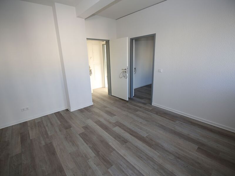 Fotografie eines geräumigen, leeren Zimmers mit mehreren Türen und Holzboden, durchdrungen von natürlichem Licht.