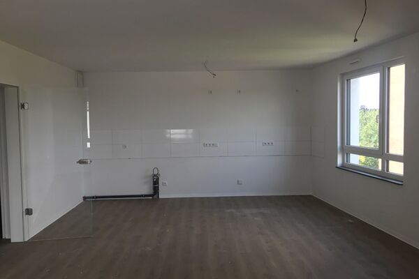 Großer, leerer Wohnraum mit vorbereiteten Anschlüssen für eine Küchenzeile und großen Fenstern, die viel Licht bieten.