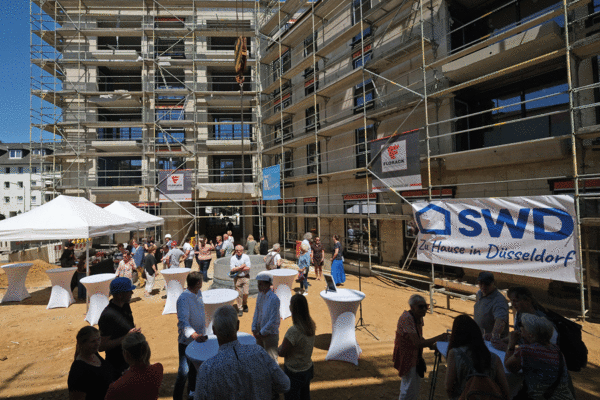 Ein Bild von einem Richtfest auf einer Baustelle mit Gästen, die sich um einen Tisch mit Erfrischungen versammeln, was einen Meilenstein im Bauprozess markiert.