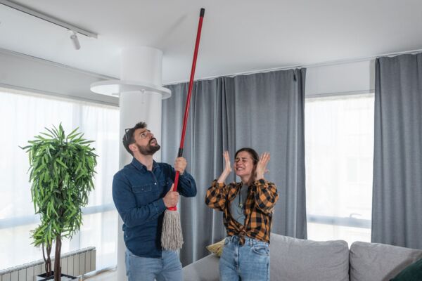 Ein Bild, das die Herausforderungen des Zusammenlebens in dicht besiedelten Wohngebieten darstellt. Es zeigt einen Mann, der einen Besen gegen die Decke haut. Während eine Frau neben ihm ihre Ohren bedeckt.